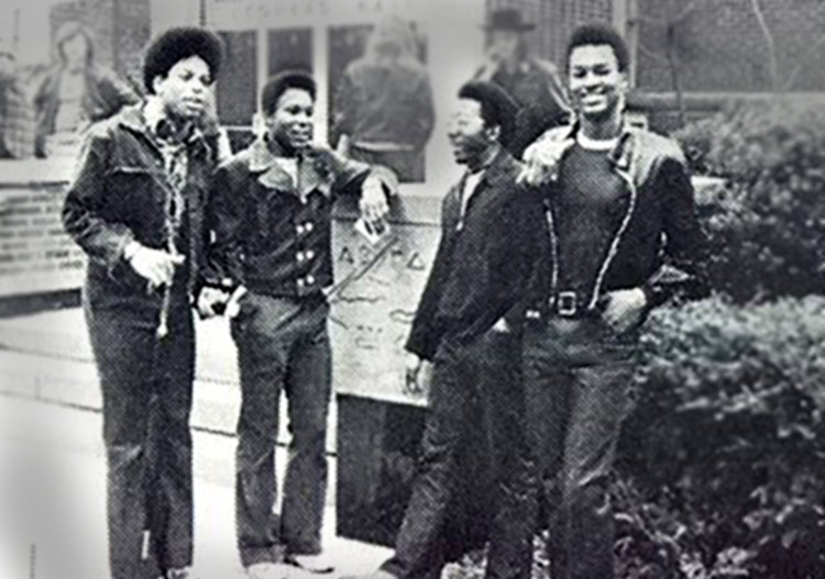 Four young men in fall clothing stand around an upright concrete slab with engraved symbols at the entrance of a building with shrubbery around it. Other people can be seen in the background.