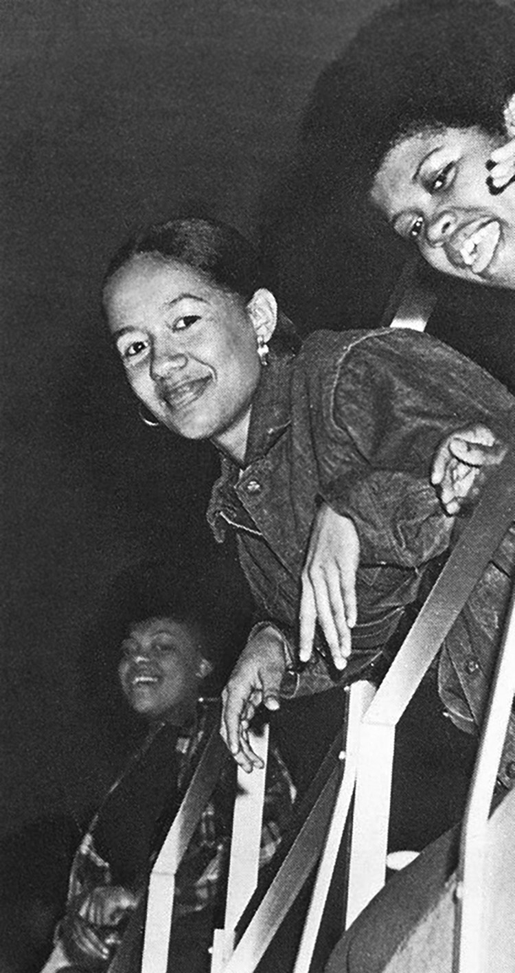 Black-and-white, indoor photograph, shot from below, of three young women leaning against a metal railing and watching something below. The woman on the right is talking, and the woman in the center is smiling and looking at the camera. 