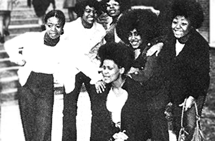 In this black-and-white photo, six young women in early-fall clothing pose outside with concrete stairs in the background. One of the six is kneeling in front, and the others have a hand on her shoulder.