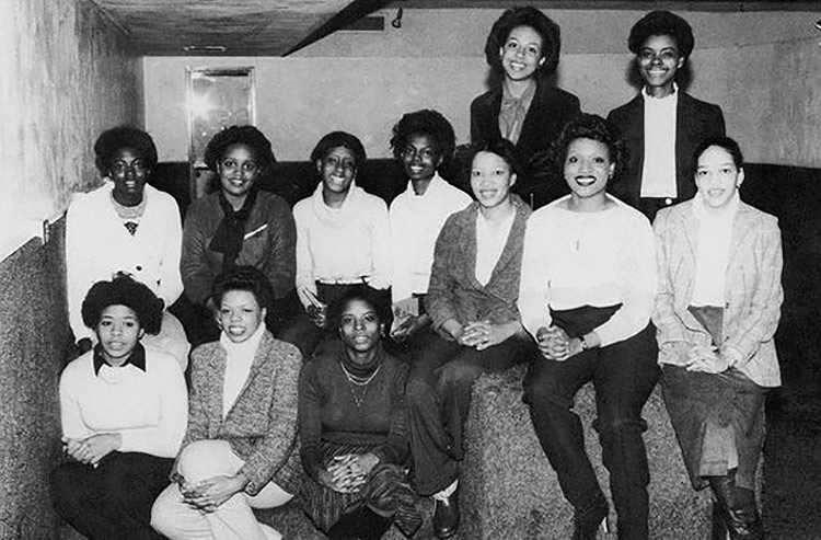 In this black-and-white photo, 10 young women in dress clothes pose on three levels. Three sit on lower stairs, seven sit one level up at the top of the stairs that appear to extend to a carpeted seat, and two appear to be standing in the back.