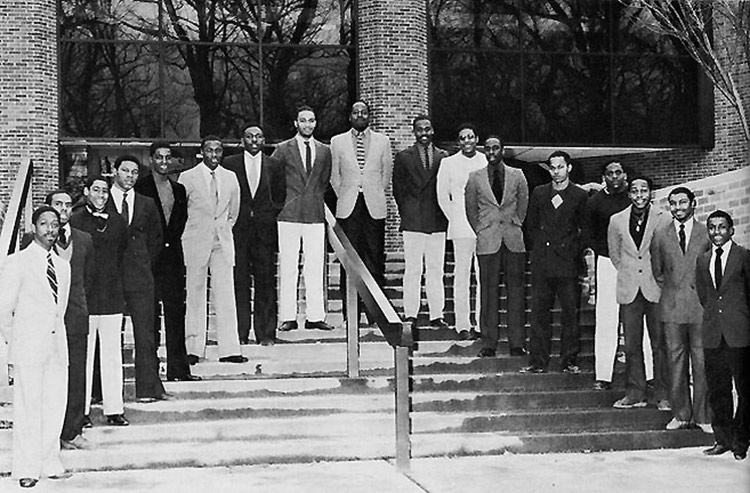In this black-and-white photo, 17 young men in coats and ties of various shades stand in a V-shape on wide, outdoor stairs, which are in front of the entrance to a building with a dark glass and brick façade. There are railings in the middle and on either side.