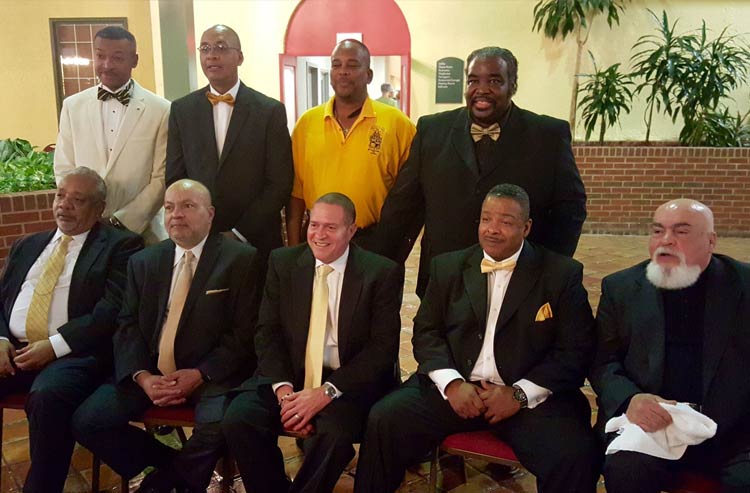 Nine older Black men—most wearing black suits and gold ties—pose in an indoor courtyard-type area with some plants in red-brick planters in the background. Five of the men are seated in front, and four stand behind them.