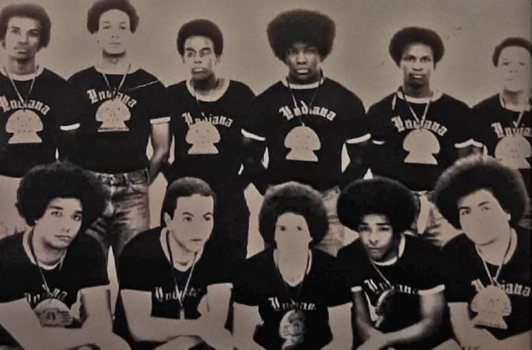 Low quality, black-and-white photo of 11 young Black men wearing black T-shirts with “Indiana” across the front and wearing large, mushroom-shaped symbols around their necks. The men are in two rows, with the front five kneeling and the back six standing.