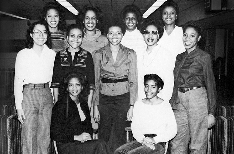 In this black-and-white photo, 11 young women in dress clothes smile and pose in a lounge-type setting with furniture with vertical stripes in the background. Two women are seated in front, five stand behind them, and four are higher in the back.