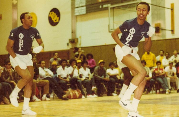 Two young men in white shorts, white gloves, and navy-blue T-shirts with the Greek symbols for Phi Beta Sigma dance in a gymnasium with an audience of young people seated behind them in folding chairs.