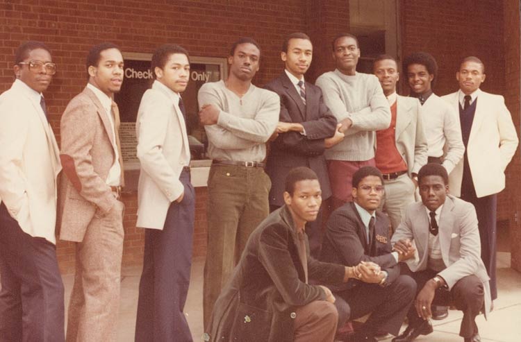 Twelve young men in dress clothes pose in front of a building. Nine men are standing in back, and three are kneeling in front. The center three in the back and all three kneeling have linked hands.