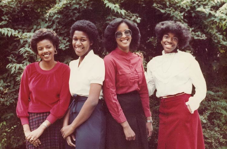 Four young women in dress clothes and styled hair pose in a row in front of trees and shrubbery.