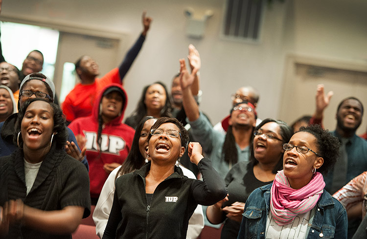 More than 20 people in casual clothing sing passionately in an indoor setting with two sets of double doors in the background. Several have their hands raised. The three women in the foreground are clear, and the other singers are more blurred the farther back they are in the group.