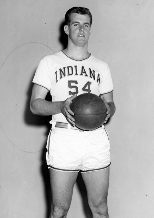 A black-and-white photo of a college-age Frank Cignetti, holding a basketball and wearing a white uniform made up of a short-sleeved shirt with "INDIANA 54" and shorts, standing in front of a plain wall