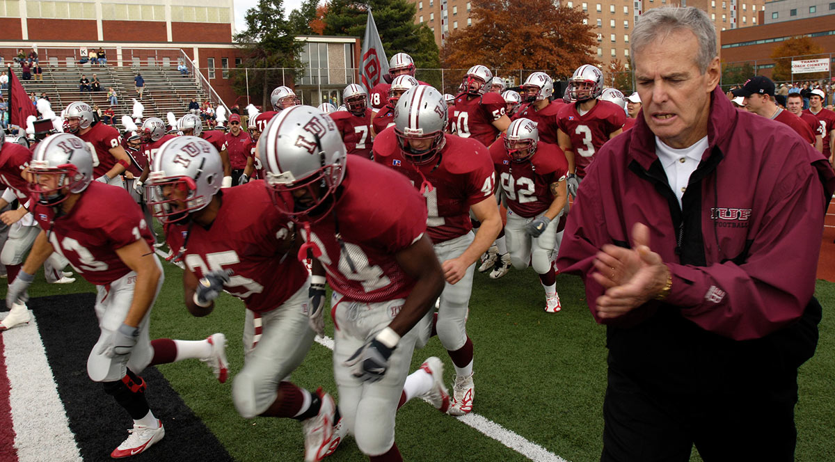 The IUP football team, in silver helmets, red jerseys, and white pants, runs onto a football field next to coach Frank Cignetti, who is clapping in a dark red IUP sweat suit.