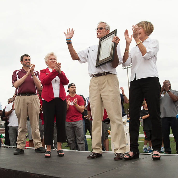 Standing on a long, black podium, Frank Cignetti holds a framed document and lifts his right hand as he speaks, flanked by a man and woman on each side of him and with several people standing behind the podium, some of them clapping.