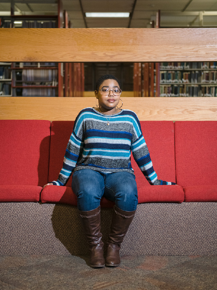A student sits on a red padded seat in the library. She's wearing brown, tall boots, jeans, and a striped blue and gray sweater. 