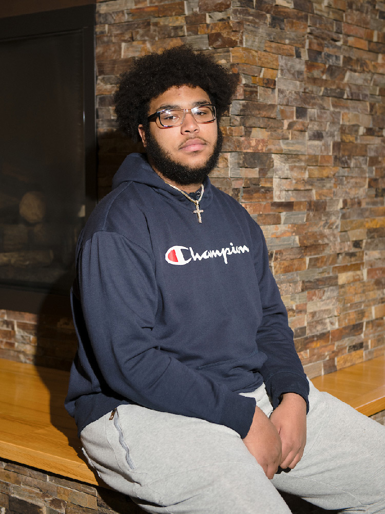 A student sits on a bench in front of a stone walled fireplace wearing gray sweatpants and a blue hoodie.