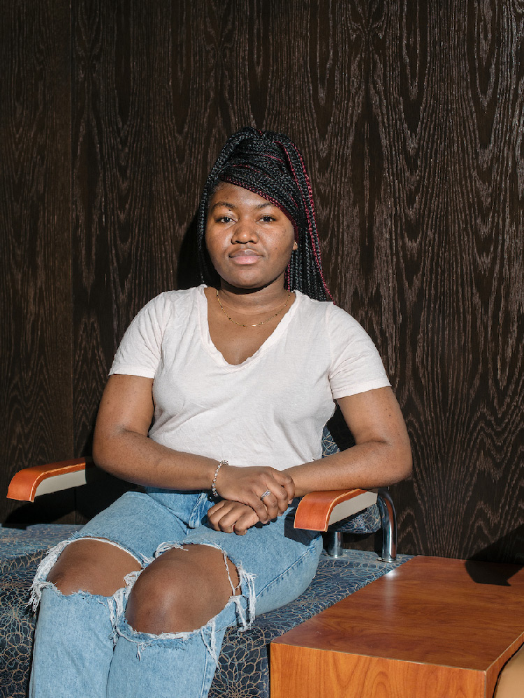 A student sits on a bench in front of a wood-paneled wall. She’s wearing jeans with holes in the knees and a white T-shirt.