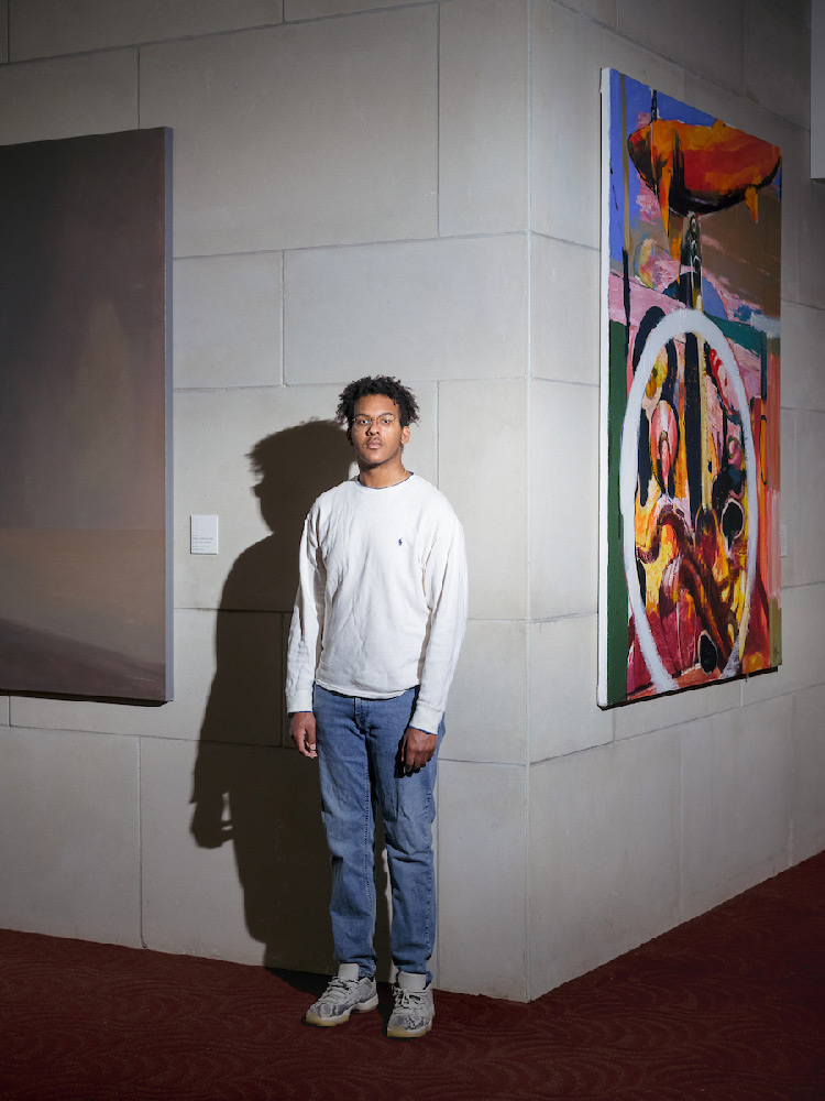 A  student wearing jeans and a white shirt stands near the corner of a wall. His shadow is seen on the wall, which has a colorful poster on it.