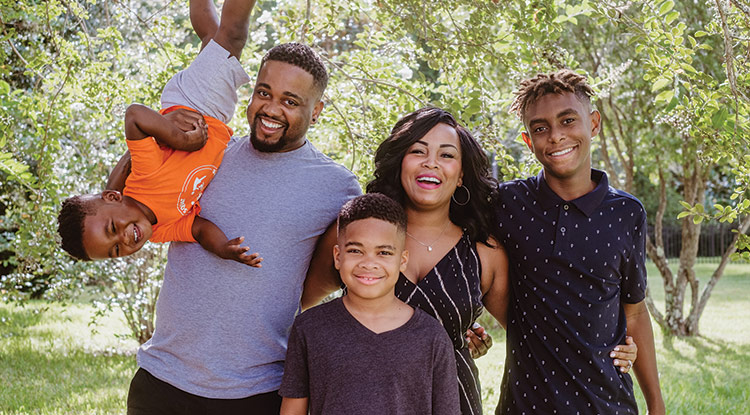 An outdoor portrait of members of the Palmer family smiling as they stand in front of some trees on a sunny day. The father, mother, and oldest son have their arms around one another’s backs, and the father is holding a toddler upside-down. The middle son is in front.