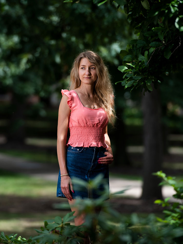 Portrait of a woman with long, blonde hair in a peach-colored shirt and a jean skirt, looking off to her right, with her left hand on her hip, surrounded by trees in a park-like setting, with a grassy area and a sidewalk visible behind her