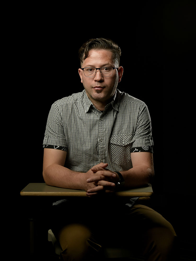 Portrait of a man with short, dark hair and glasses, wearing a black and white checkered shirt and tan pants, sitting on a chair with a built-in desk, folding his hands on top, and looking into the camera. His left side is partly shadowed, and he is in front of a black background.