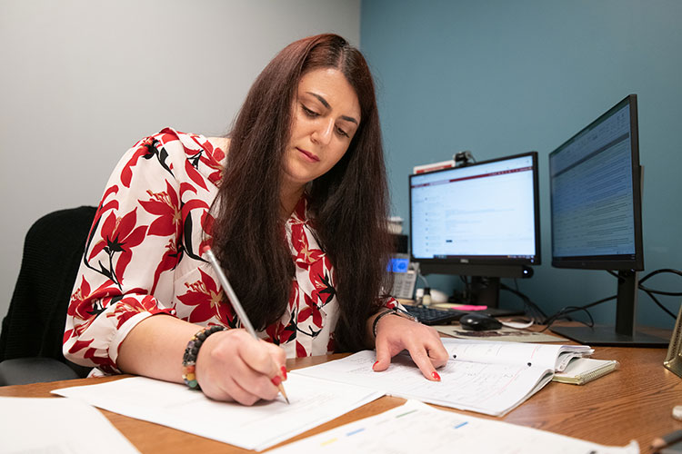 A woman with long, dark hair works at an office desk, where she is holding a notebook open with one hand and writing on a piece of paper with the other. A computer keyboard, mouse, and two large monitors are on the desk in the background.