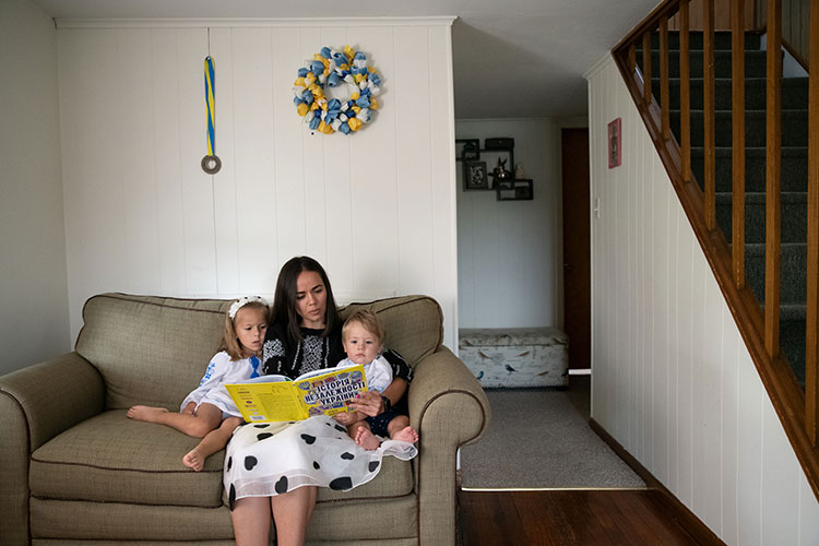 A woman sits on a small, tan couch, reading a children’s book with a yellow cover and words in Ukrainian to a little girl on her right and a toddler boy on her left. A hallway and stairs are visible on the right, and a medal with a blue and yellow ribbon and a blue and yellow wreath hang on the wall above the couch.