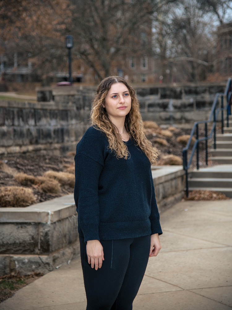 Outdoor portrait of Claudia Pauley standing in a courtyard area with a low stone wall behind her, a taller stone wall behind that, and a building in the background