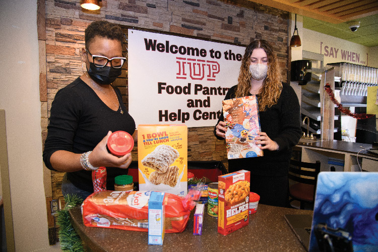 Malaika Turner, left, and Claudia Pauley hold and examine grocery items while standing behind a counter with more groceries. Both are masked, and a sign reading, “Welcome to the IUP Food Pantry and Help Center” is hanging on a wall behind them.