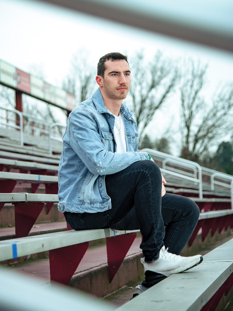Portrait of a serious-looking Connor Kelly sitting in empty stadium seating with one foot on a metal bench and his arm rested on his knee
