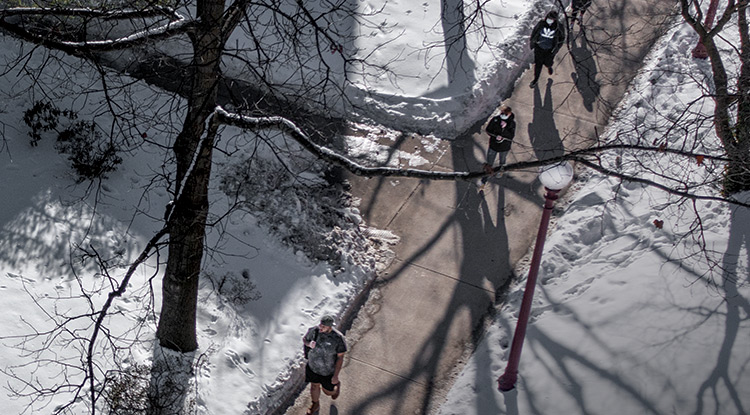 View from above of six students, some wearing backpacks, walking on a cleared sidewalk beneath bare trees on a sunny day with perhaps a foot of snow on the ground 
