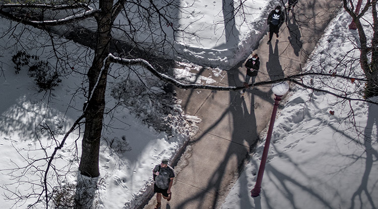 View from above of six students, some wearing backpacks, walking on a cleared sidewalk beneath bare trees on a sunny day with perhaps a foot of snow on the ground