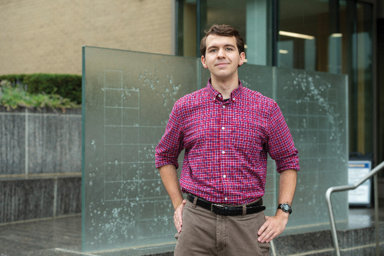 Portrait of a slightly smiling Noah Garrett standing with his hands on his hips in front of a vertical frosted-glass grid in front of a building