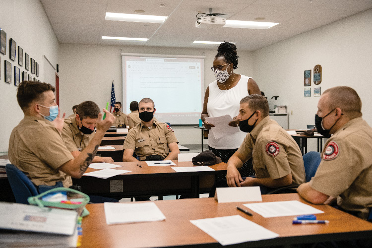 Veronica Watson stands, holding papers, in a classroom setting and talks with a group of five seated police cadets wearing matching tan, collared shirts. All are wearing masks, and another small group of cadets is visible behind them.