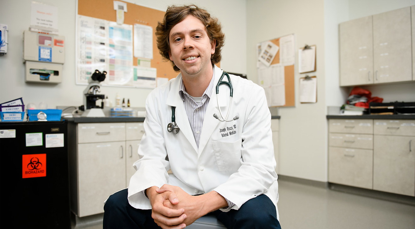 A young man wearing a buttoned, white lab coat over business casual clothing and a stethoscope around his neck sits on a chair in the middle of a medical exam room or lab with a microscope in the background. He smiles for the camera as he leans slightly forward with his forearms resting on his legs and his hands clasped in front of him.