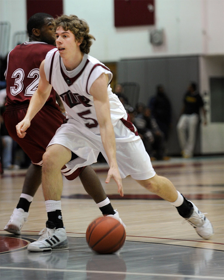 A young man in a white IUP jersey dribbles a basketball on a court during a game with an opposing player in a dark-red jersey behind him and his back to the player with the ball. Spectators standing against a wall are visible in the background.