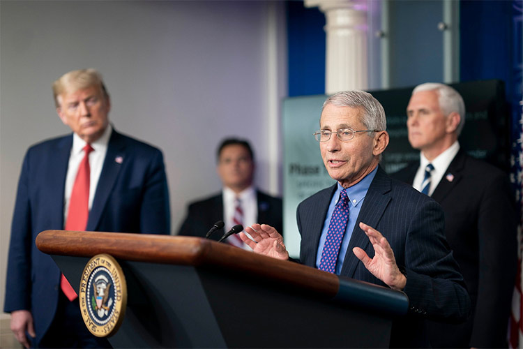 An older man wearing a suit and glasses stands at a podium with a presidential seal on its front and with his hands raised slightly in front of him as he talks. Three men wearing suits and listening to him are slightly blurred in the background.