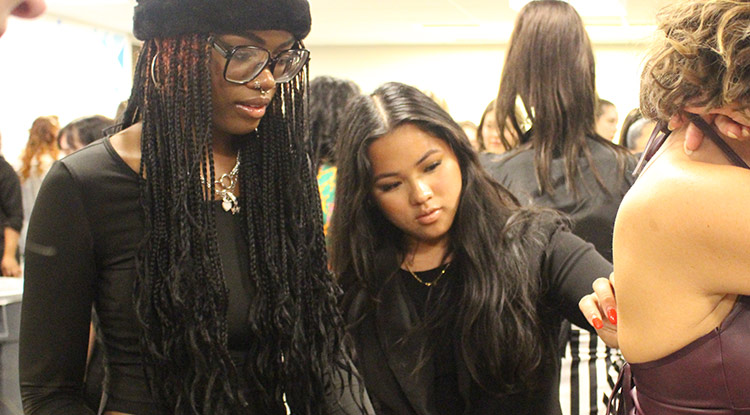 Two female college students who are helping backstage at a fashion show help a model with something that needs to be adjusted at the back of her shirt.