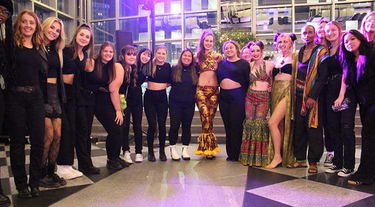 A group of about 18 college-age students, mostly women, form a U-shaped group as they pose for a group photo.