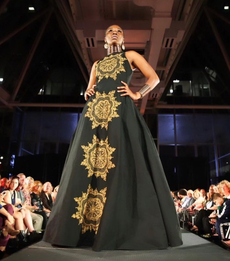 A model, Jordan Brown, walks on a raised runway during a fashion show wearing a long, dark gown with four, large, gold, sun-like emblems sewn in a row going down the front. The audience is seated below along the runway.