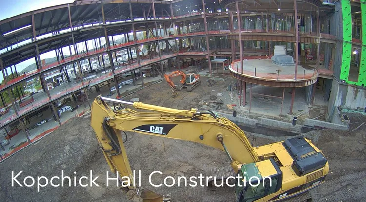 Heavy machinery working in the dirt while a half constructed building sits in the background.  The words "Kopchick Hall Construction" is on the bottom left.
