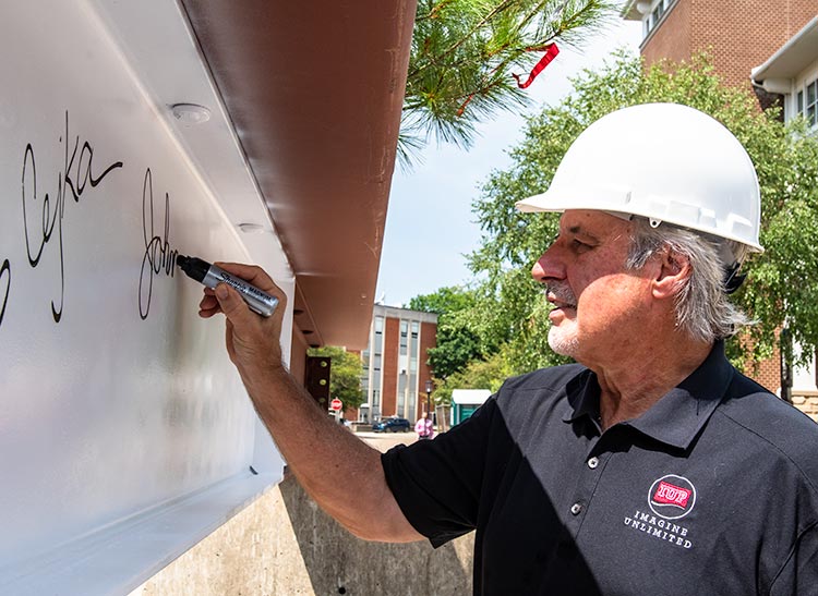 Dr. John Kopchick in a hard hat signing his name on an I-beam