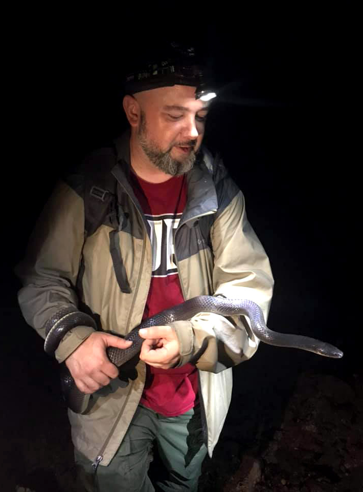 Dr. Townsend standing in the dark with a head mounted flashlight holding a snake