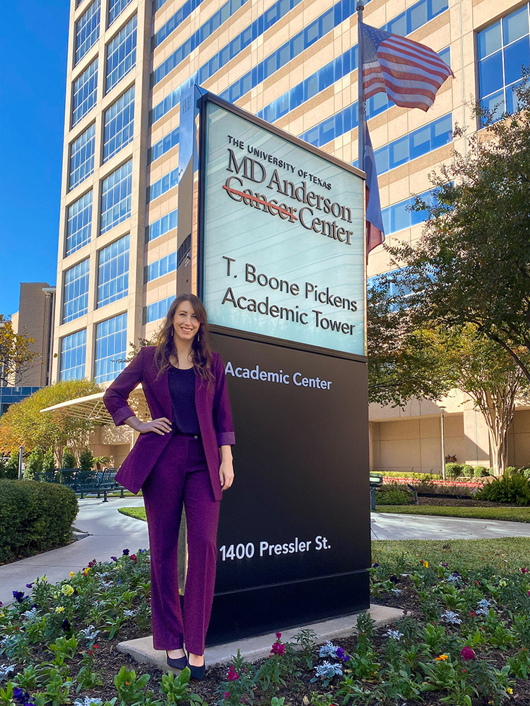 A woman with long hair stands by the sign for the University of Texas MD Anderson Cancer Center in Houston, Texas. There are flowers planted around the sign. The building stands many stories high behind her with blue sky around it.