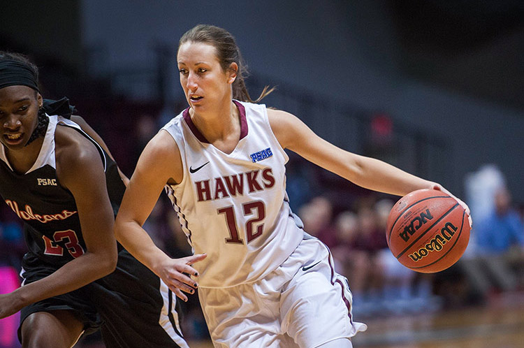 In a basketball game, Leslie Stapleton, number 12, wearing a white uniform, has the ball and is moving fast around an opposing player wearing black.