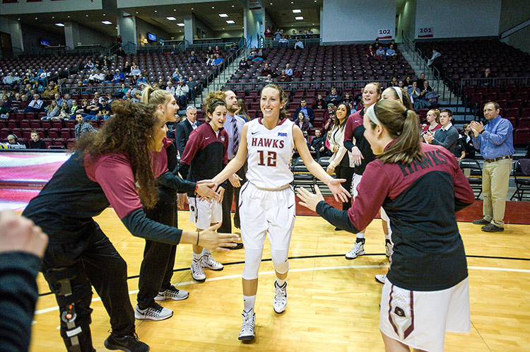 It's a break in a basketball game or after the game, with people in the stands. Leslie Stapleton, in a white basketball uniform with her hair pulled back, is congratulated by teammates, as she goes through the center of two lines of players, mostly wearing crimson and black jackets over their uniforms, who are giving her high-fives from both lines.
