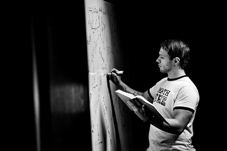 Vincent Thompson holds a text book open as he writes mathematical equations on a white board across a wall, with darkness framing the black and white image.