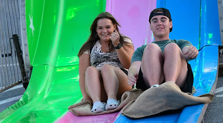 Two students riding down a colorful carnival slide on burlap bags