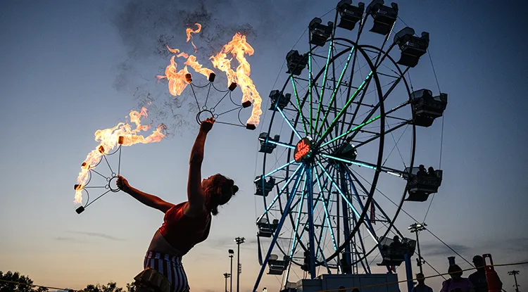A performer holding flaming sticks in front of a lit up ferris wheel