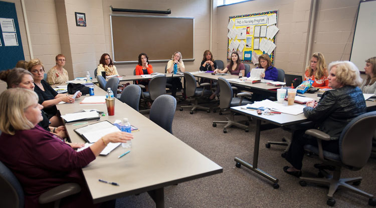 Several women of various ages, with notebooks and papers in front of them, have a discussion as they sit in a classroom in a U-shape made of long tables.