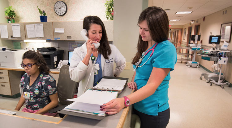 Three women in medical clothing are at a nurses’ station in a hospital setting. Inside the station, one in dark scrubs is sitting and typing, with her computer off camera, and one in a white coat is standing and talking on the phone while looking at a chart that a woman in blue scrubs on the opposite side of the counter is also looking at. Medical equipment is lining the hallway in the background.