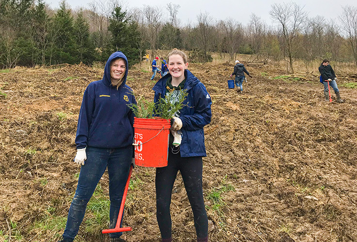 Shelby Zakrzewski, left, holding a planter, and Kaitlin Knopsnider, holding up a bucket of saplings, smile as other team members are planting behind them in a field with trees in the background.