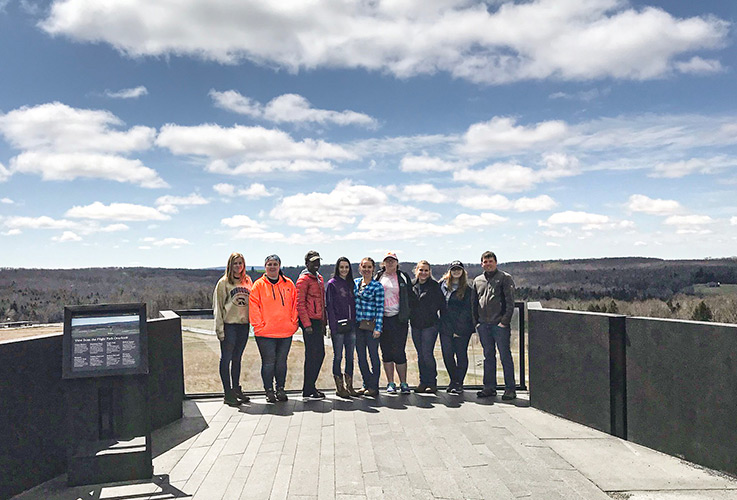 Eight students and faculty member Michael Tyree on the right stand on a deck at the Flight 93 memorial, with the site and its surrounding landscape in the background.
